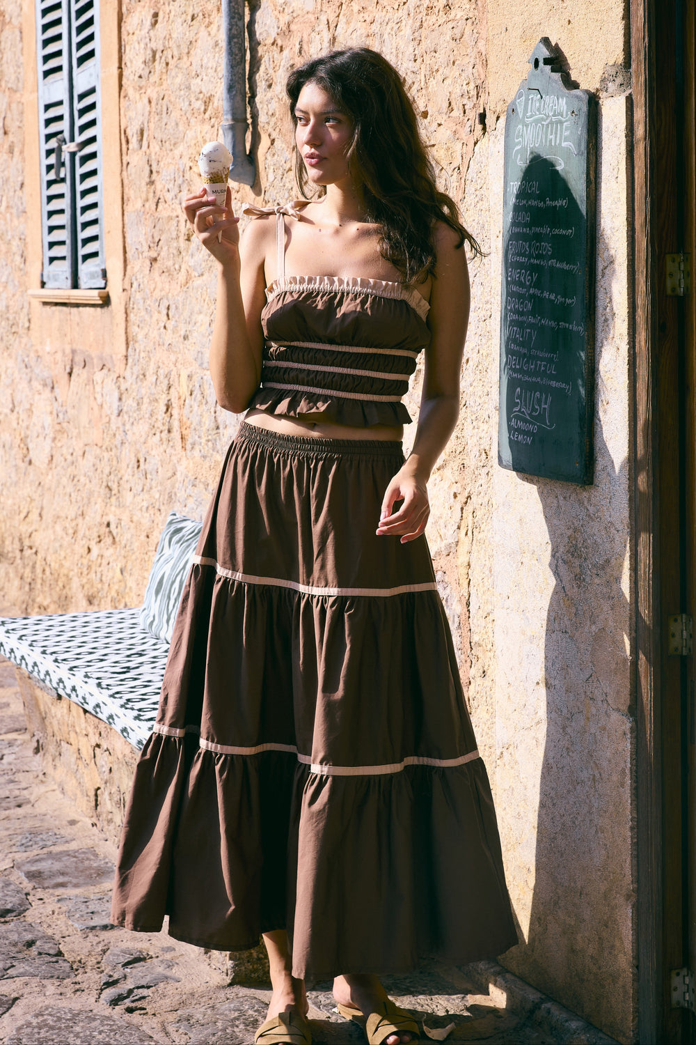 Woman in a long dress linen standing against a stone wall in a sunlit street.